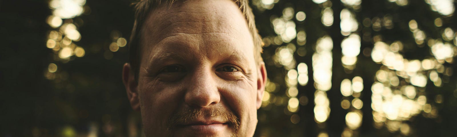 A man with short, tousled hair and a goatee smiles gently at the camera, standing outdoors in front of a blurred background of trees and soft evening light.