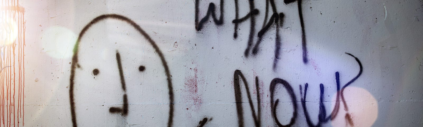 Graffiti with staid face and “What Now?” spray painted on the the wall of an abandoned building.