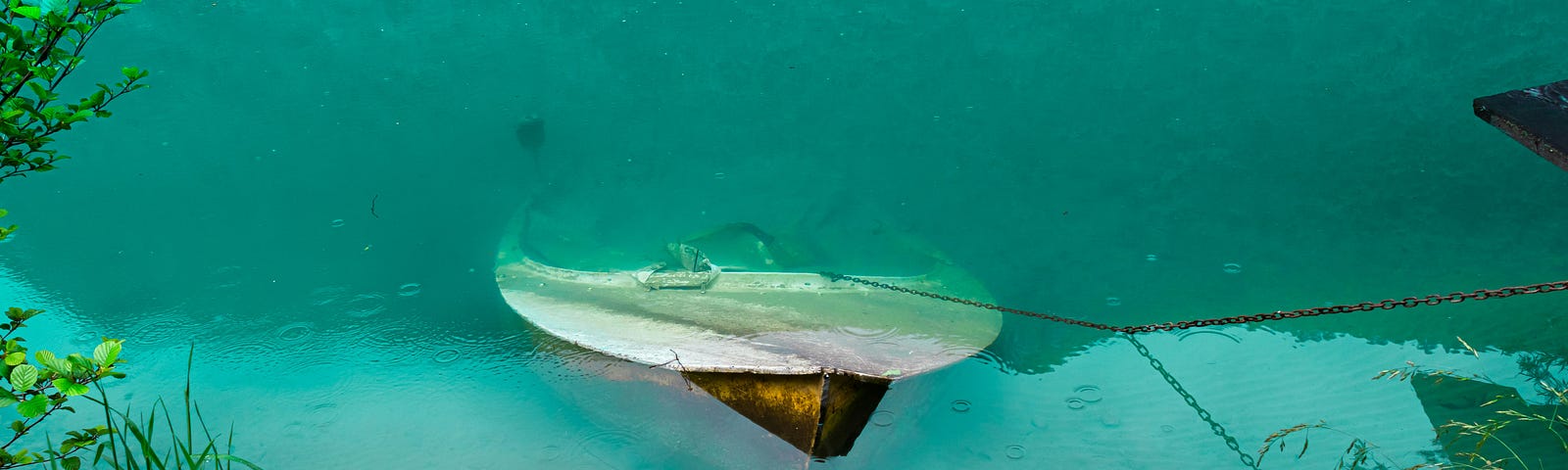 The front edge of a submerged boat is poking out of crystal clear water. You can count pebbles on the bottom of the lake. Trees and foliage reflected back in deep greens, lush and vibrant.