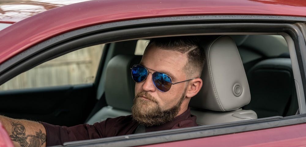 A man in obnoxious sunglasses stares with a gimlet eye from the window of a car.