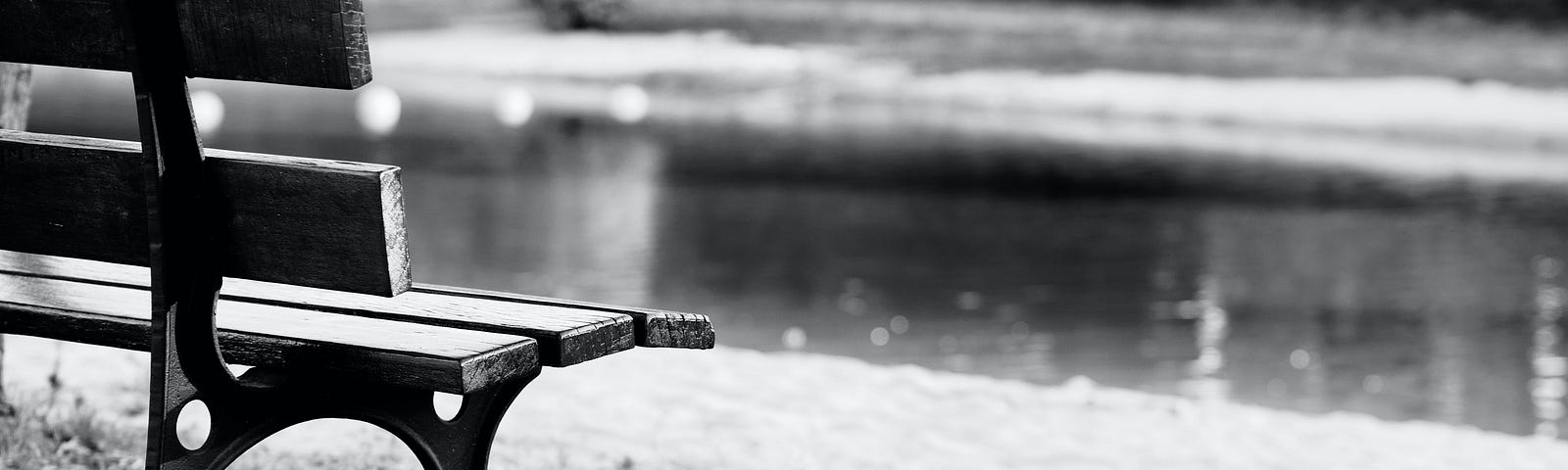 A black and white photo of an empty bench next to a pond.