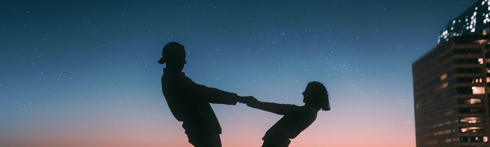 Silhouette of a couple on a nighttime rooftop, holding hands and leaning back from each other; she looks as though she may fall if he lets go.