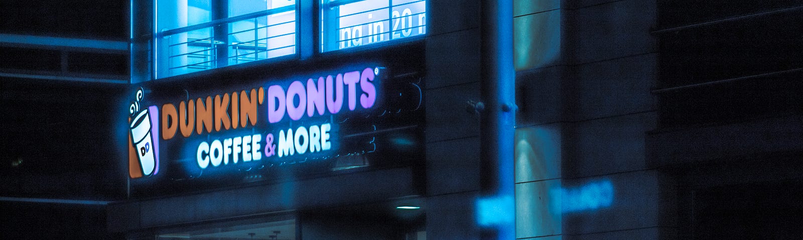 Nighttime photograph of Dunkin’ Donuts restaurant exterior, with glowing in the dark above the windows of the restaurant, and a brightly lit office space on the 2nd floor above.