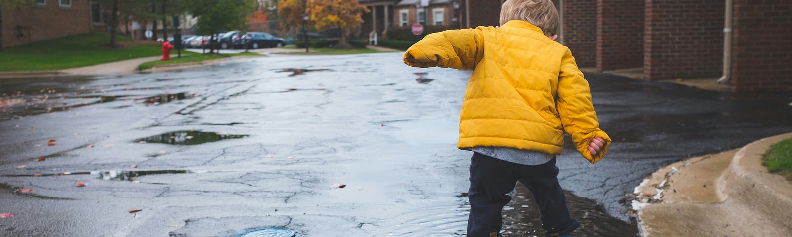 A small child wearing a yellow coat jumping into a puddle on an empty street.