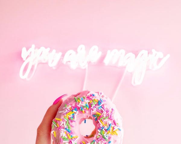 A hand holding a donut with “you are magic” in neon on the wall in the background