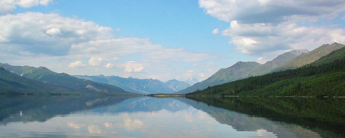 View of a lake from a boat or canoe. There are mountains in the background and a partly cloudy sky above.