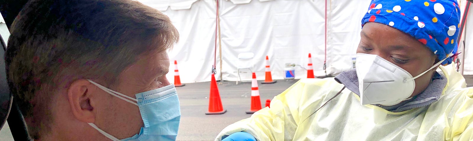 A white man wearing a salmon colored shirt in a car and wearing a blue surgical mask is receiving a vaccine through the window from a Black nurse wearing scrubs, blue gloves, an N95 masks, and a blue handkerchief with polka dots.