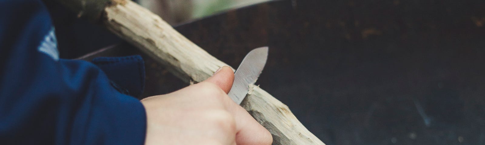 An image of someone holding a small knife with a sharp tip whittling off the bark of a wooden stick.
