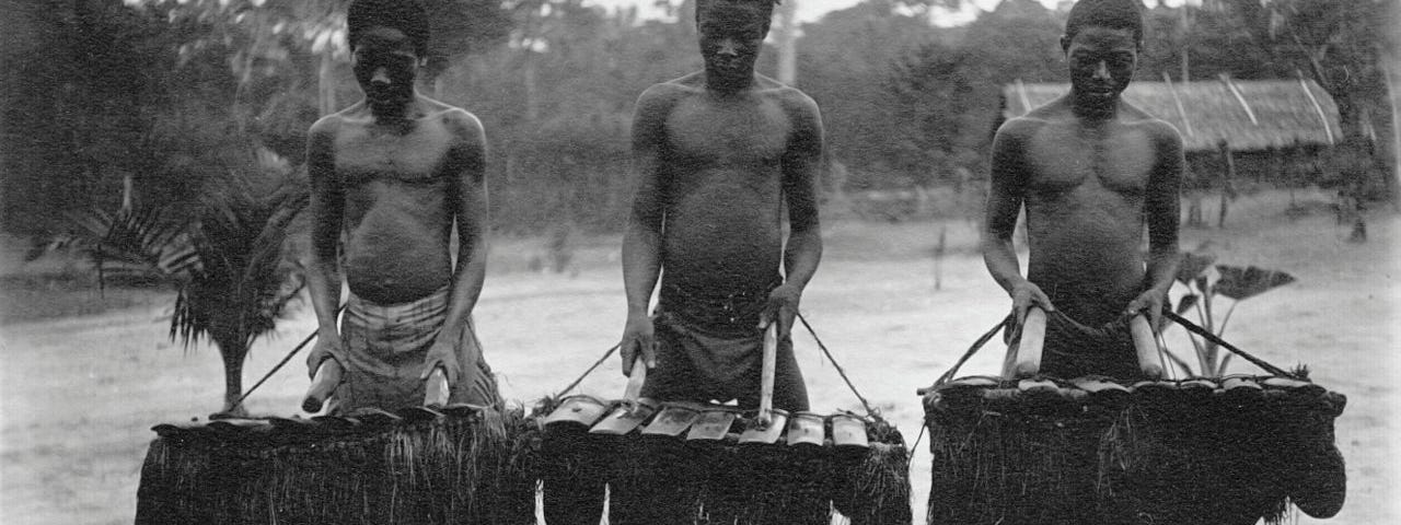 Fotografia em preto e branco de três homens negros tocando instrumentos musicais.