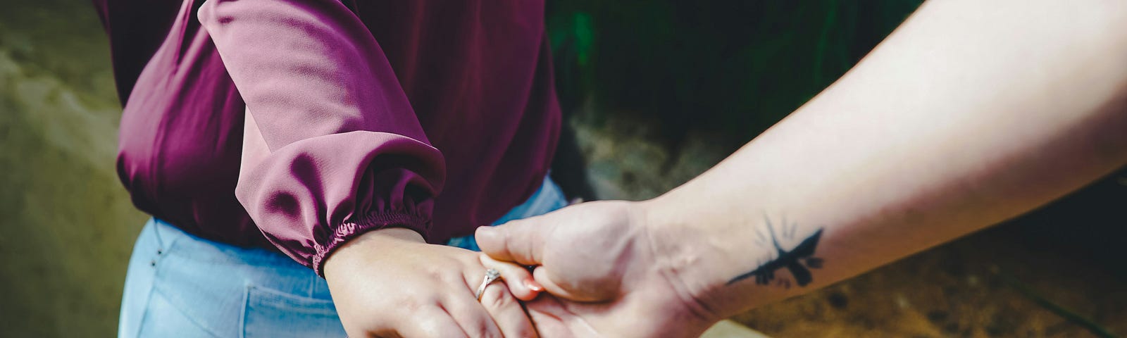 A woman wearing a wedding ring her hand behind her back, leads a man behind her along a narrow walk. His fingers are tucked into her hand. Only his arm is showing and it is bare with a small star like tattoo just above his inner wrist.