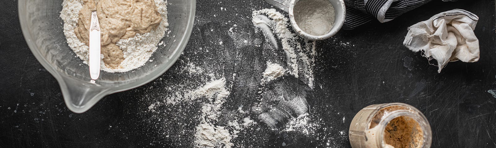 A jar of sourdough starter and a bowl of flour and starter mixture sit on a black flour dusted counter top.