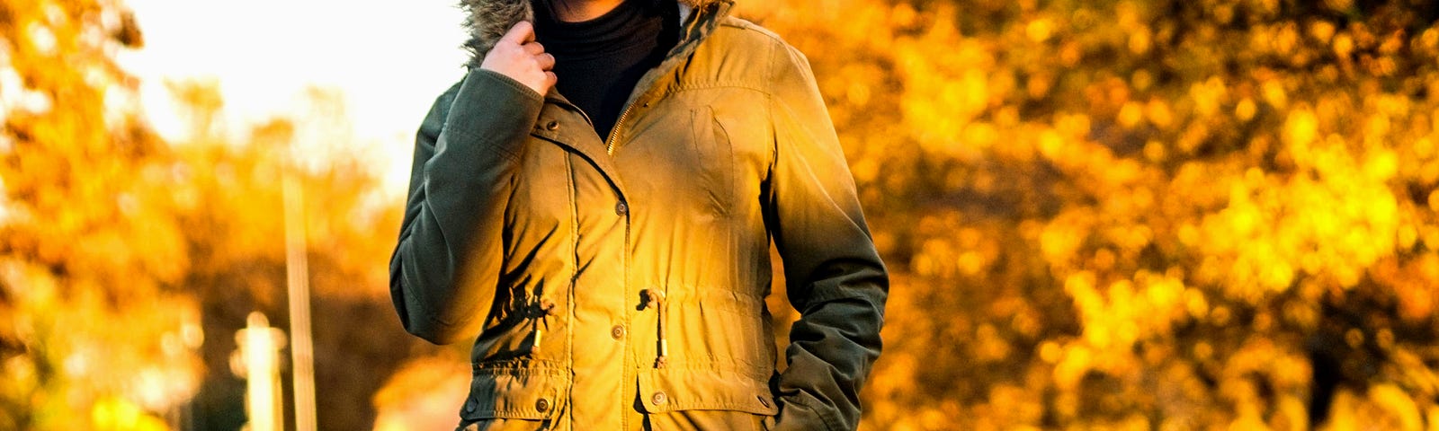 Photo of a young woman walking along a railway track, wearing red sneakers with white laces, and black leggings, and a mustard brown winter coat with fur-lined hood. Her hand is up at her hood, and she could be touching the fur lining or her hair beneath the hood. The background is lined with trees, leaves in autumn shades. The ground beneath the train tracks is large crushed gravel.