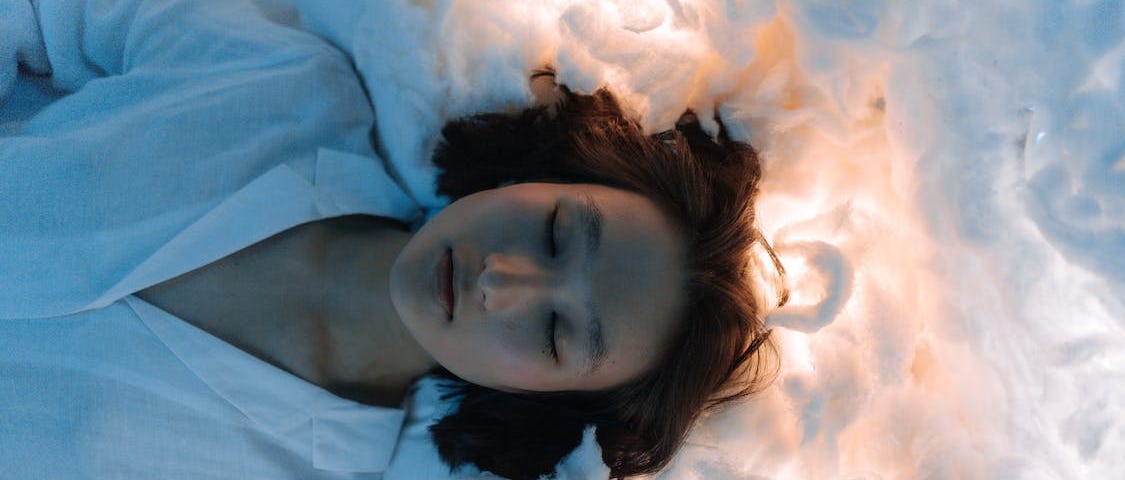 Brunette woman ina white blouse lying on white cotton with a light shining through it. Her eyes are closed and she looks deep in thought