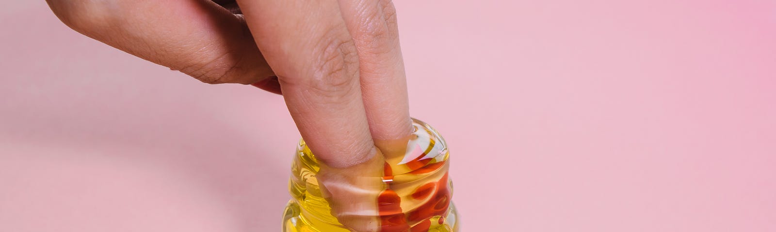 A man dips two fingers into a small jar of honey. Pink background. Honey has antibacterial and anti-fungal properties.