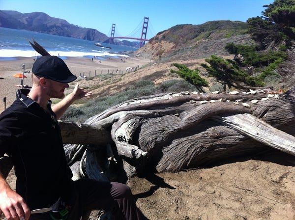 A sunny beach in San Francisco with the iconic Golden Gate Bridge in the background. The bridge is actually red, though. It was named for the straight it spans over the ocean. A man wearing a feathered hat sits on driftwood, gesturing towards a row of oyster shells.