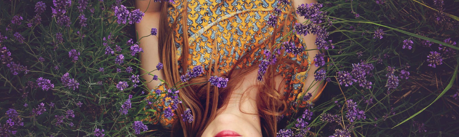A red-haired woman in a gold and green printed top lays serenely, with eyes closed, on a bed of purple and green lavender-like plants.