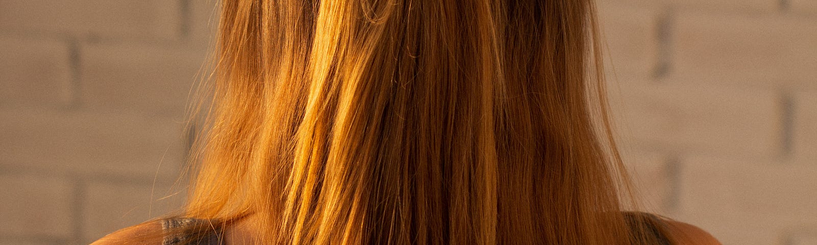 The back of the head of a woman with long reddish hair, standing straight, shoulders back, facing a whit brick wall.