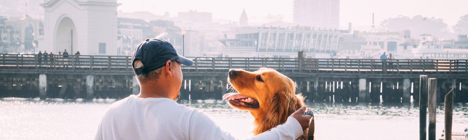 Man standing with large golden retriever looking him in the face, with a pier and harbor buildings in the background.