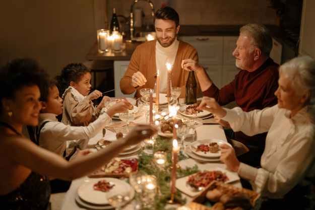 Christmas dinner table with a white millennial man sat at the top of the table flanked by an old white couple on his right and two brown children on his left. On the other side of the chlidren there is a millennial brown woman lighting the candles on the table. The millennial man and woman smile.