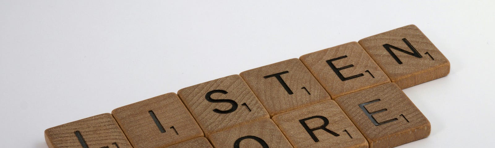 Wooden Scrabble tiles arranged to spell ‘LISTEN MORE’ on a white background.