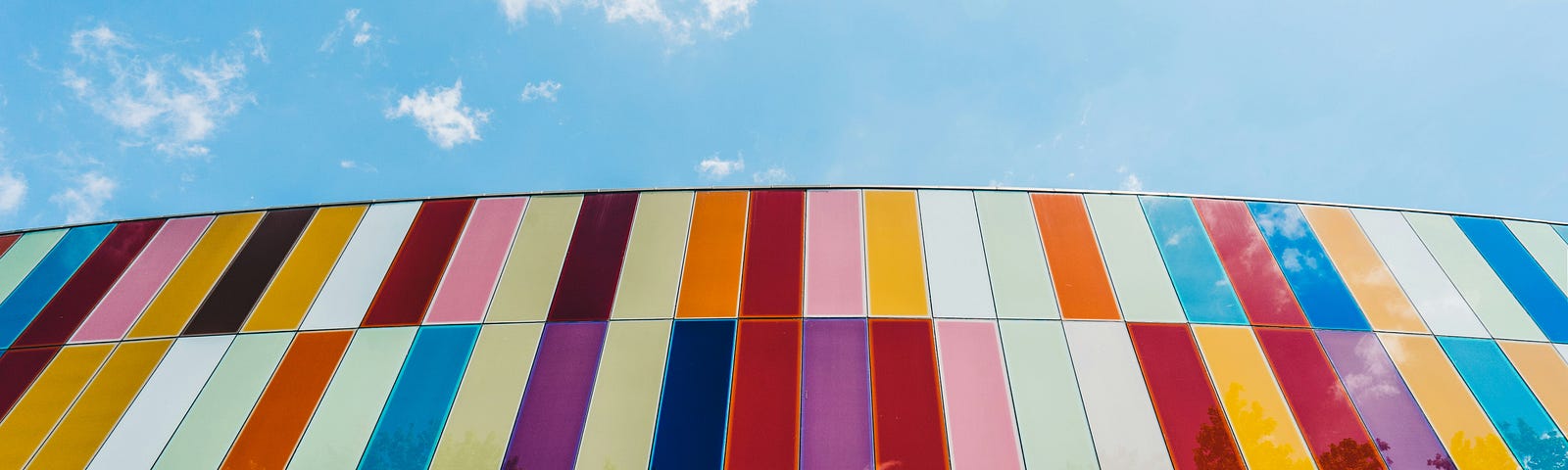 Photo of a wall with vertical rectangles placed next to each other of different colors, forming a colorful wall. Above it, you can see a blue sky with some clouds.