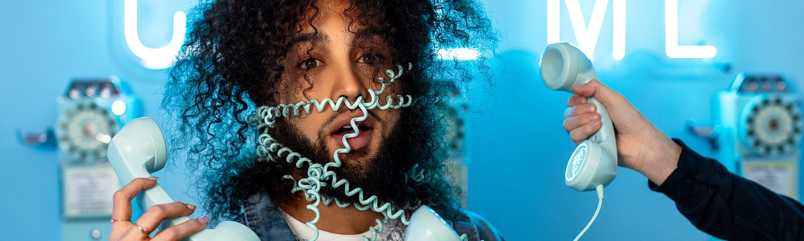 A man looking disheveled, wrapped with multiple coiling phone cords over his body and face, with multiple hands holding the phone receivers, reaching toward his face. The background is a blue neon lit sign that says “CALL ME”