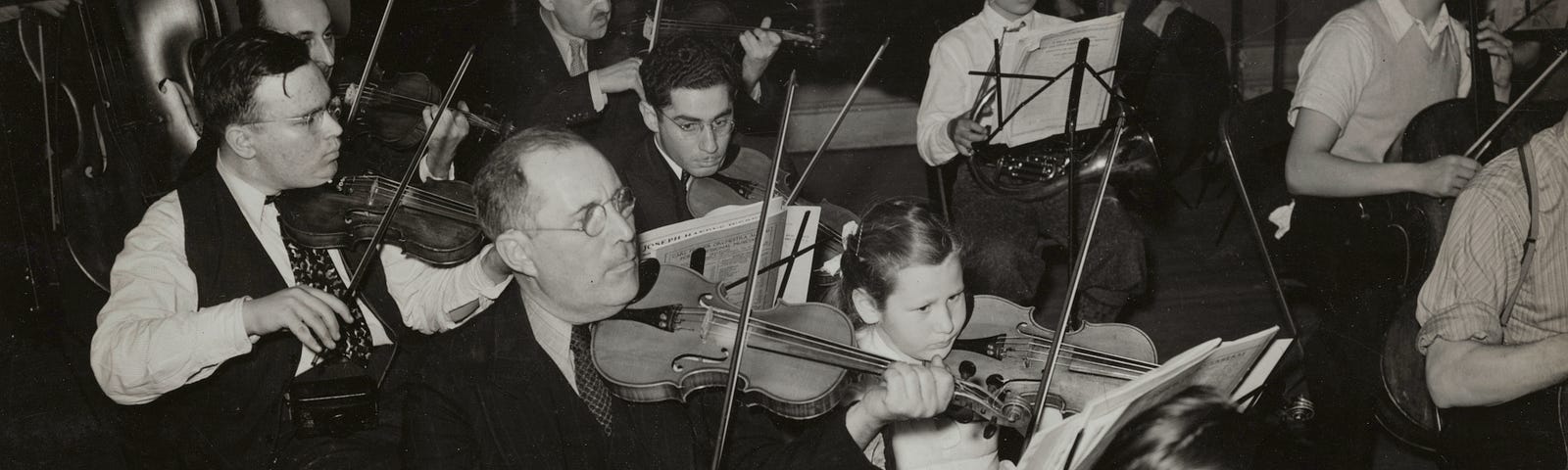 a portion of an orchestra of both adults and children mostly playing violins with a piano in the background.