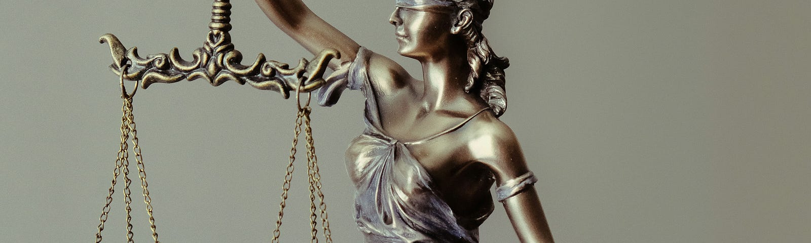 A bronze sculpture of Lady Justice. She is blindfolded, holding a sword facing down with her left hand and an even balance with her right hand. She is barefoot. Her right foot is on a small platform, and her right knee is bent. She is wearing a loose-fitting dress.