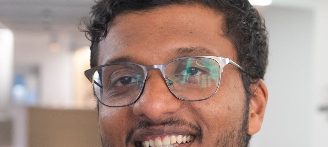 Person wearing a gray shirt and steel-framed glasses smiling at the camera. Out of focus background that looks like an office