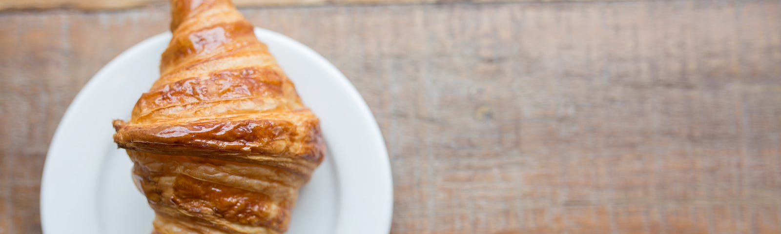 A croissant on a small white plate on a a wooden counter top.