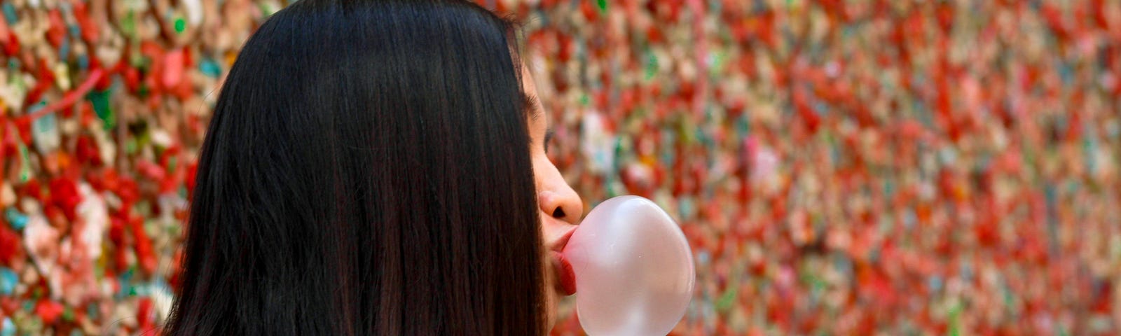 Profile of a young woman blowing a large pink bubble from chewing gum. The Seattle’ Pike Place Market gum wall is in the background.