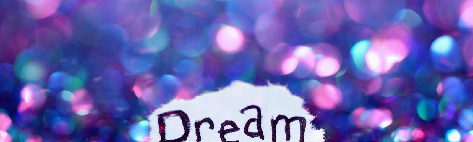 The word dream is written on a small piece of paper. It lies on a bed of tiny sparkling crystals, ranging in colour from greens over blues to purple tones.