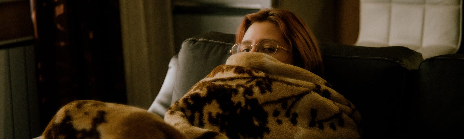 A woman is curled up on a couch underneath a blanker with her eyes peering out the top.