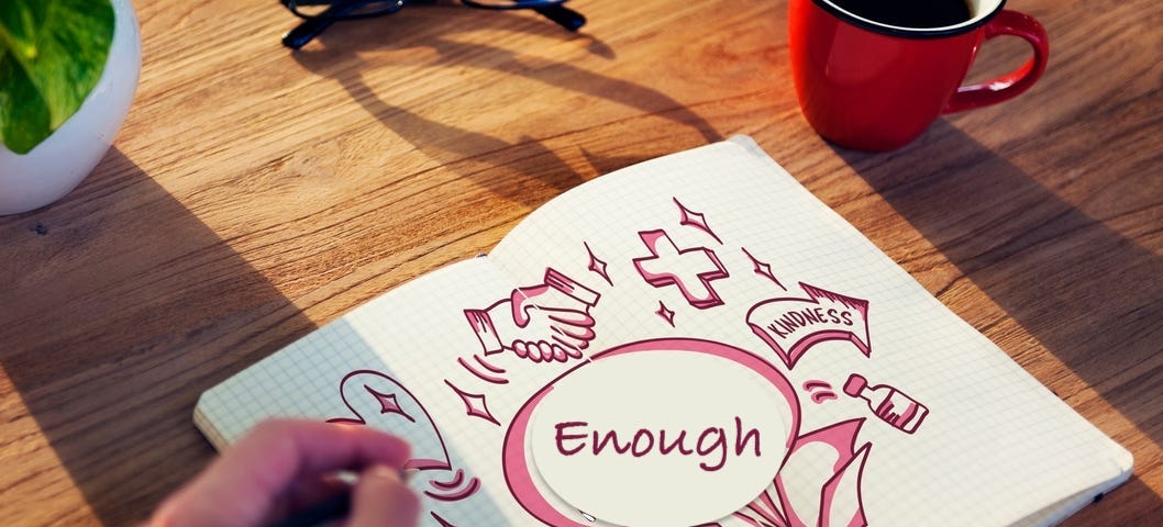 On a wooden surface, a book with jottings in red says ‘Enough’, a hand with a pen in the foreground, a red mug with black coffee in the background