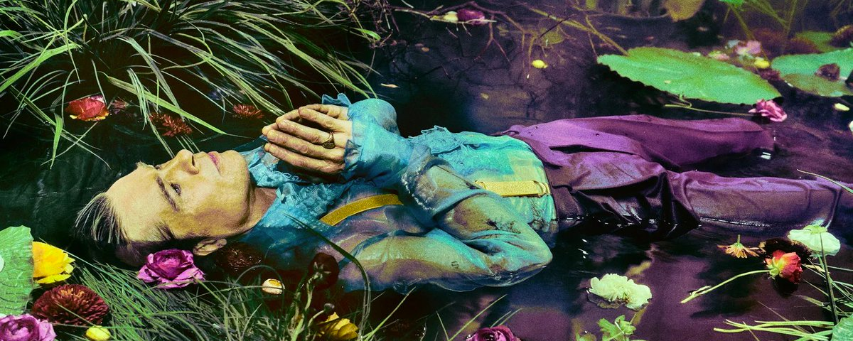 Brad Pitt lays in swampy waters face up with his hands over his chest as if in prayer. He wears a sheer blue short and purple trousers which match the purple flowers and green foliage growing in the water.