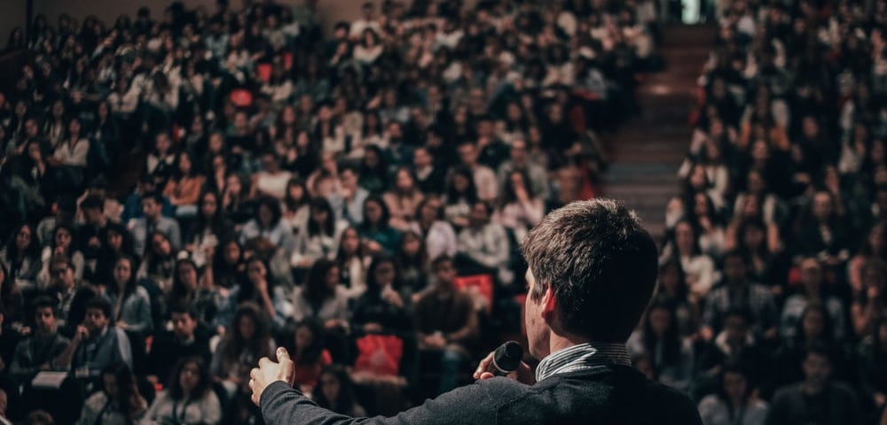 Young man speaking into a microphone in front of a crowded auditorium.