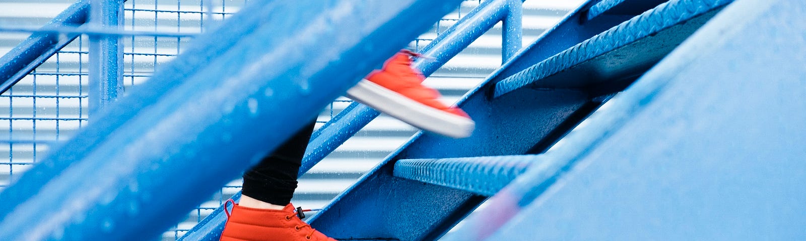 A person walks up a flight of blue-coloered stairs. The image is cropped such that we see only the person from knee level down to her red tennis shoes (trainers). Americans took fewer steps during the COVID-19 pandemic peak, and we have not gotten back to our pre-coronavirus numbers. Today we explore a worrisome trend in steps taken, especially among young individuals.