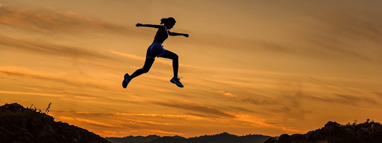 Young Woman In Sport Apparel Jumping Over A Cliff With Sunset In The Background.