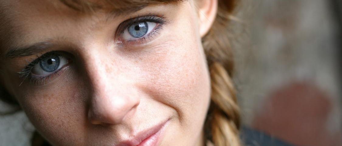 Close up of a woman with blue eyes looking into the camera with a look of quiet confidence. Her hair hangs to the left of her head in a plait, with a faded brickwall in the background