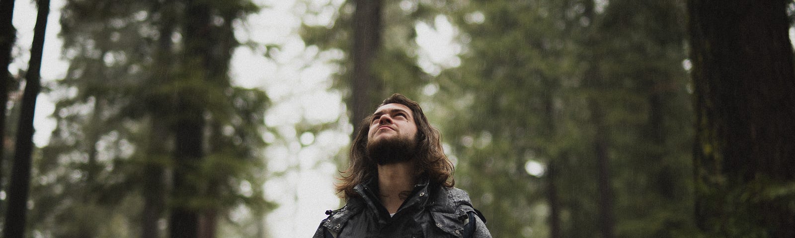 Photo of teen boy looking upward in a forest, long, dark hair, hands in pockets of coat.