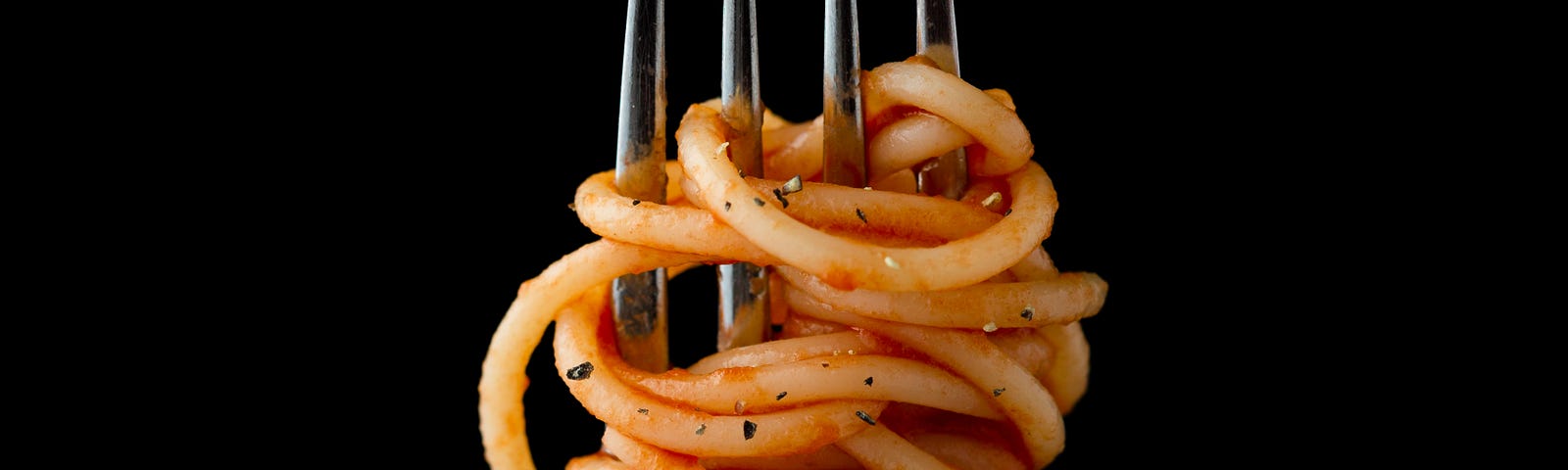 A silver fork extends upwards in the center of the image, with spaghetti weaving in and out of the tines. There are smidges of tomato sauce and spices.