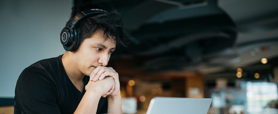 Photo of a man wearing over-ear headphones sitting at a table with his hands clasped together. He’s staring at an open laptop in front of him.