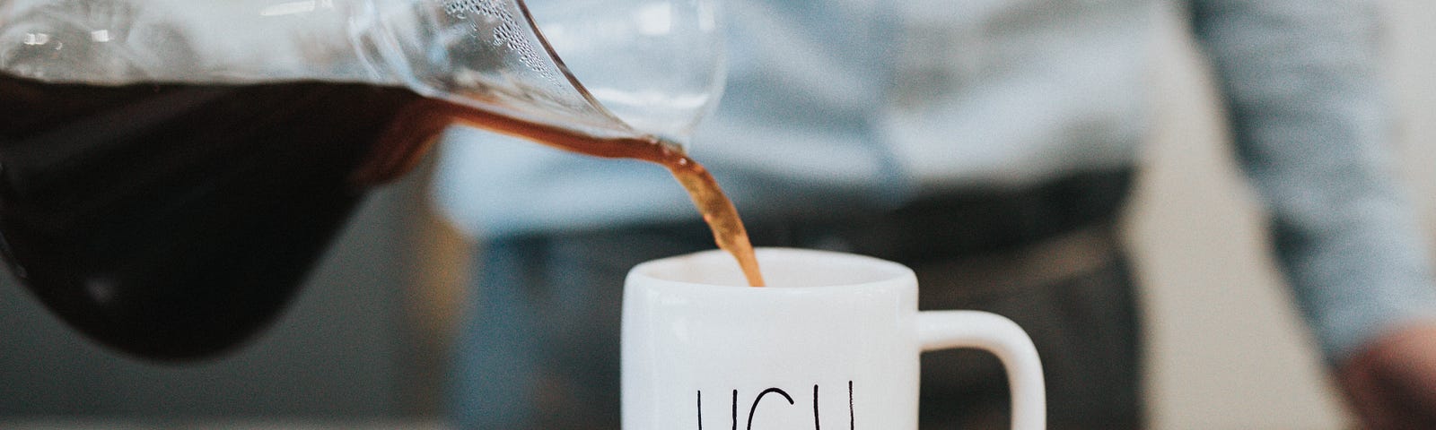 Photo of coffee being poured into a white mug with “UGH” written on it.