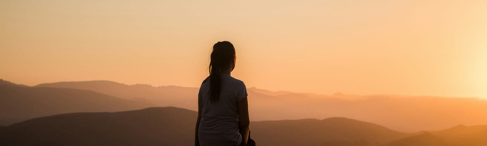The silhouette of a woman sitting on a bench looking across a range of mountains in the sunset