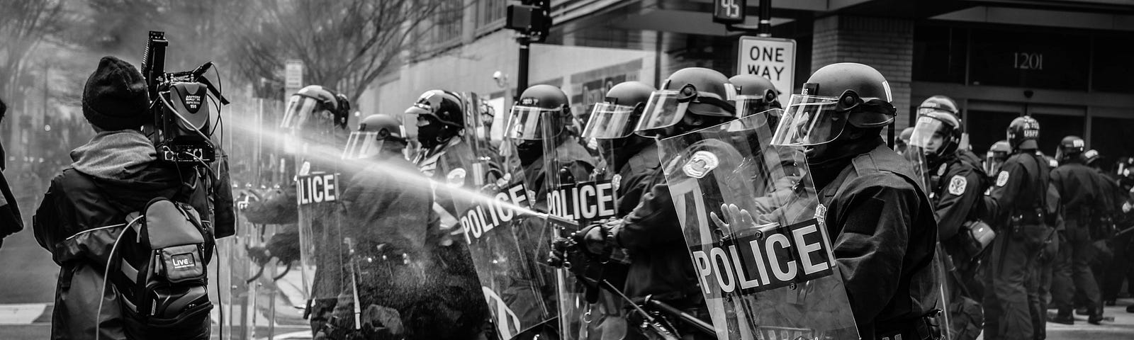 A black and white photo of riot police standing in a line across a pedetrial crosswalk; one policeman holding the equipment with the visible spraying of liquid spewing up in a concentrated stream towards presumed protestors that are just outside the view of the photo. We see the back of one camera crew member standing just in front this line of police.