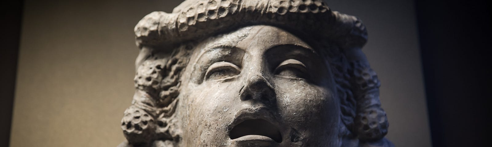 A greyish stone head statue of presumed Medusa with her mouth ajar and hair in more likeable braids (no visible snakes).