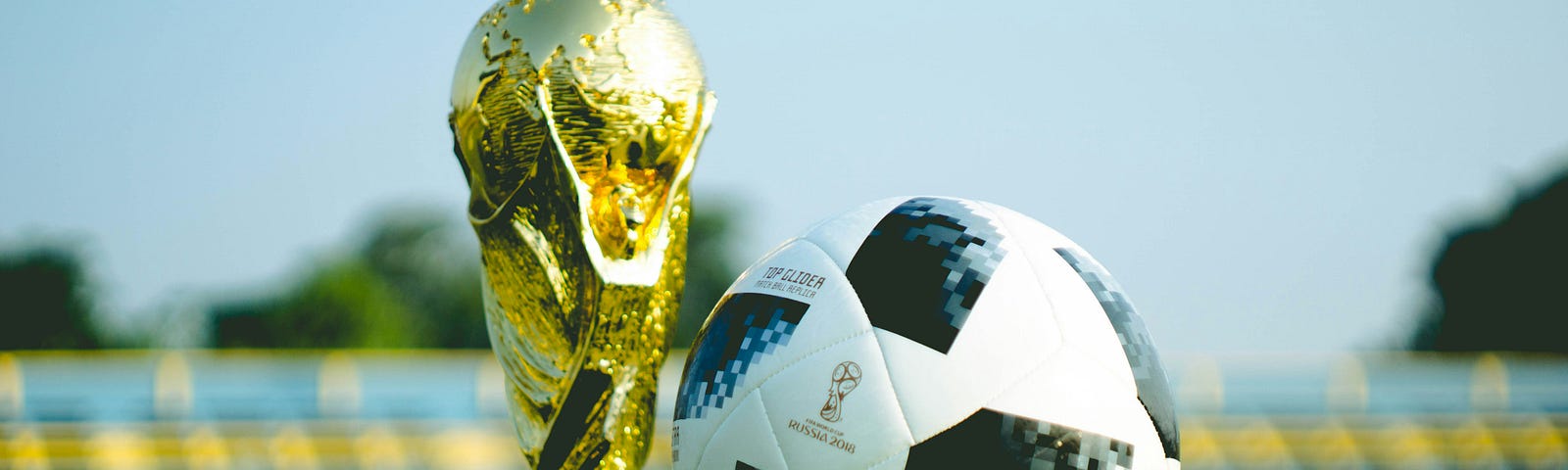Football and trophy in close-up on an outdoor pitch with stands behind.