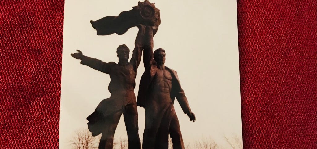 The photo is of two men standing together and holding up a giant medallion with a large ribbon waving toward the left. There are bare trees in the bottom of the background.