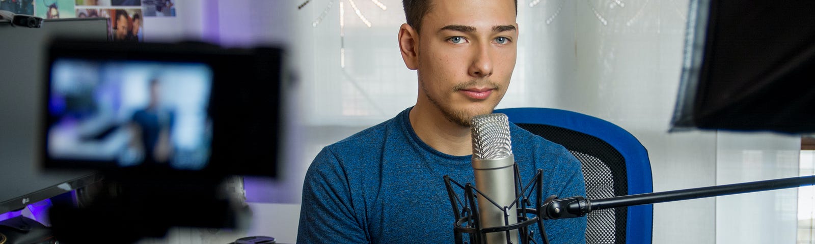 A white man with slicked back brown hair sits behind a filming set-up with a silver microphone propped up near his mouth on the left and a camera aimed towards his face on the right. He is wearing a cerulean blue t-shirt, has short facial hair, and majestic eyebrows. He is looking at the photographer with a somber expression.