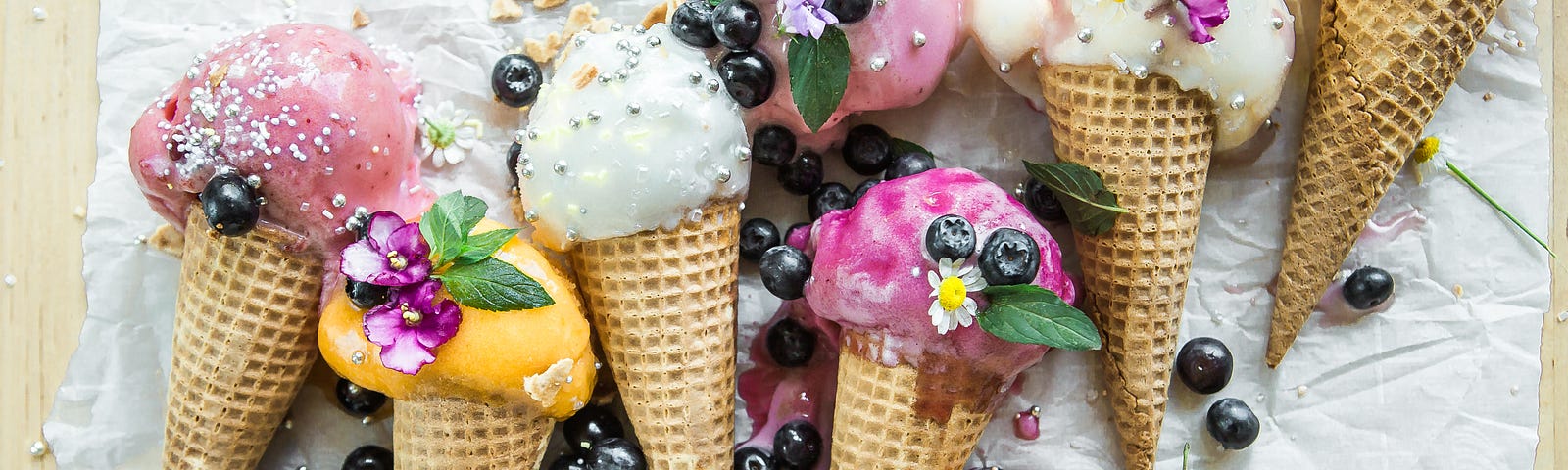 Many sugar cone of various flavors of ice cream with blueberries and mint leaves sprinkled around